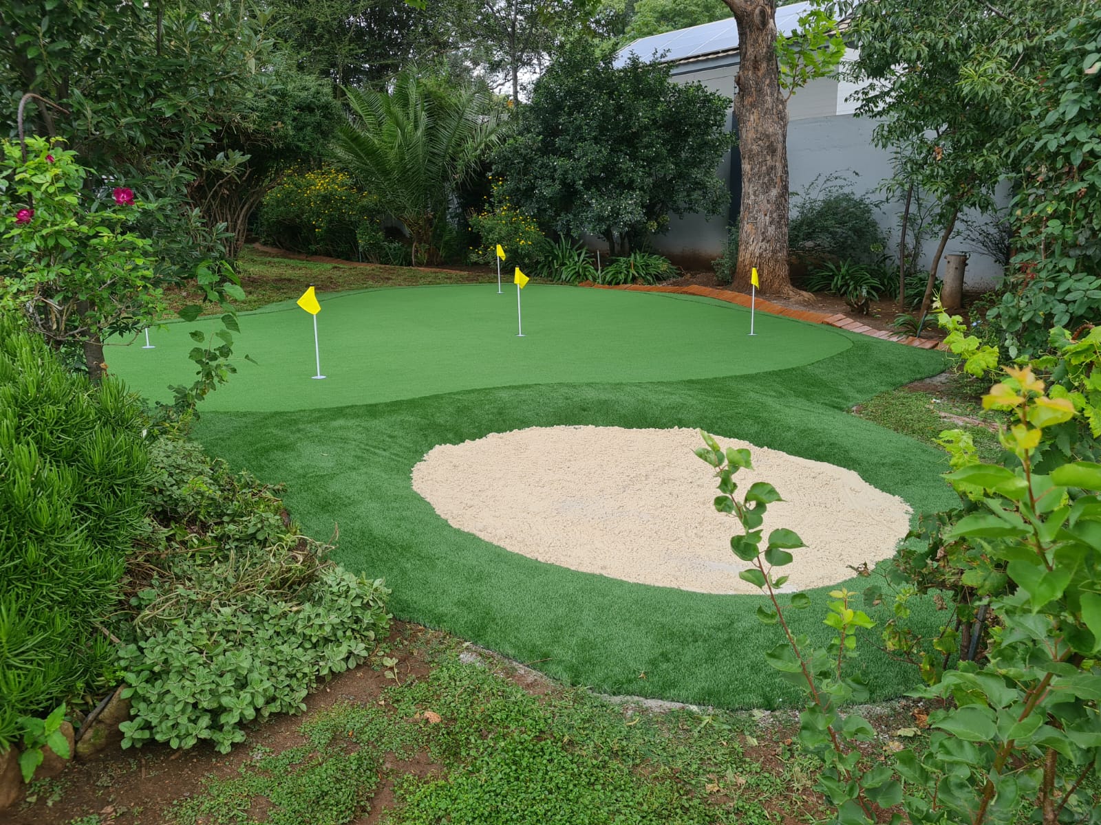 Long outdoor synthetic putting green with multiple cup flags installed along a residential building side-yard in South Africa.