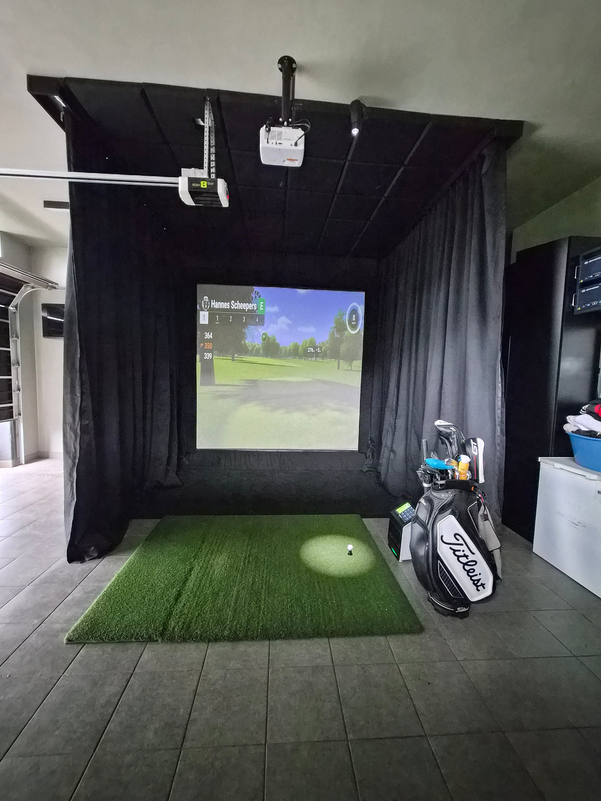 Single-car garage golf simulator conversion featuring a black curtain enclosure, hitting mat, and floor-mounted launch monitor.