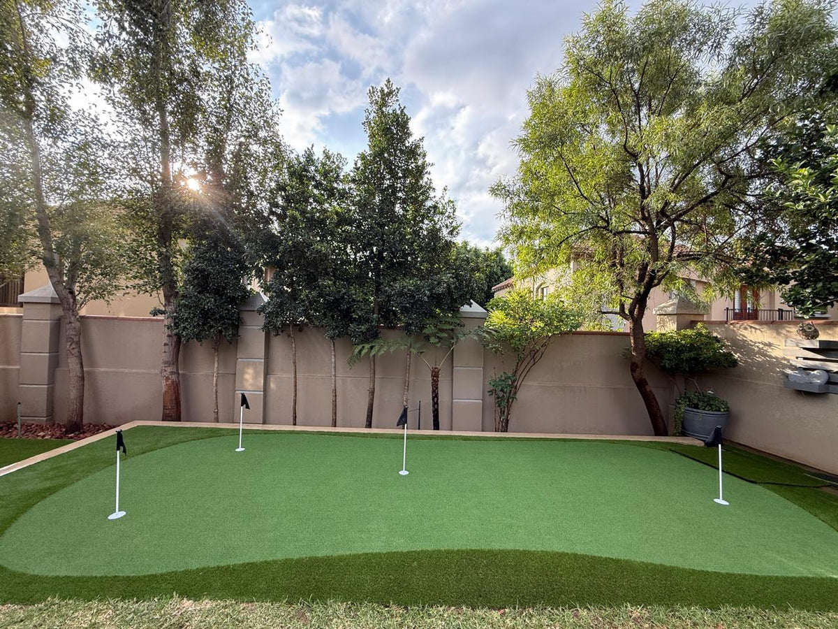 Custom backyard artificial turf putting green installation featuring four pin flags surrounded by natural landscaping and garden trees.