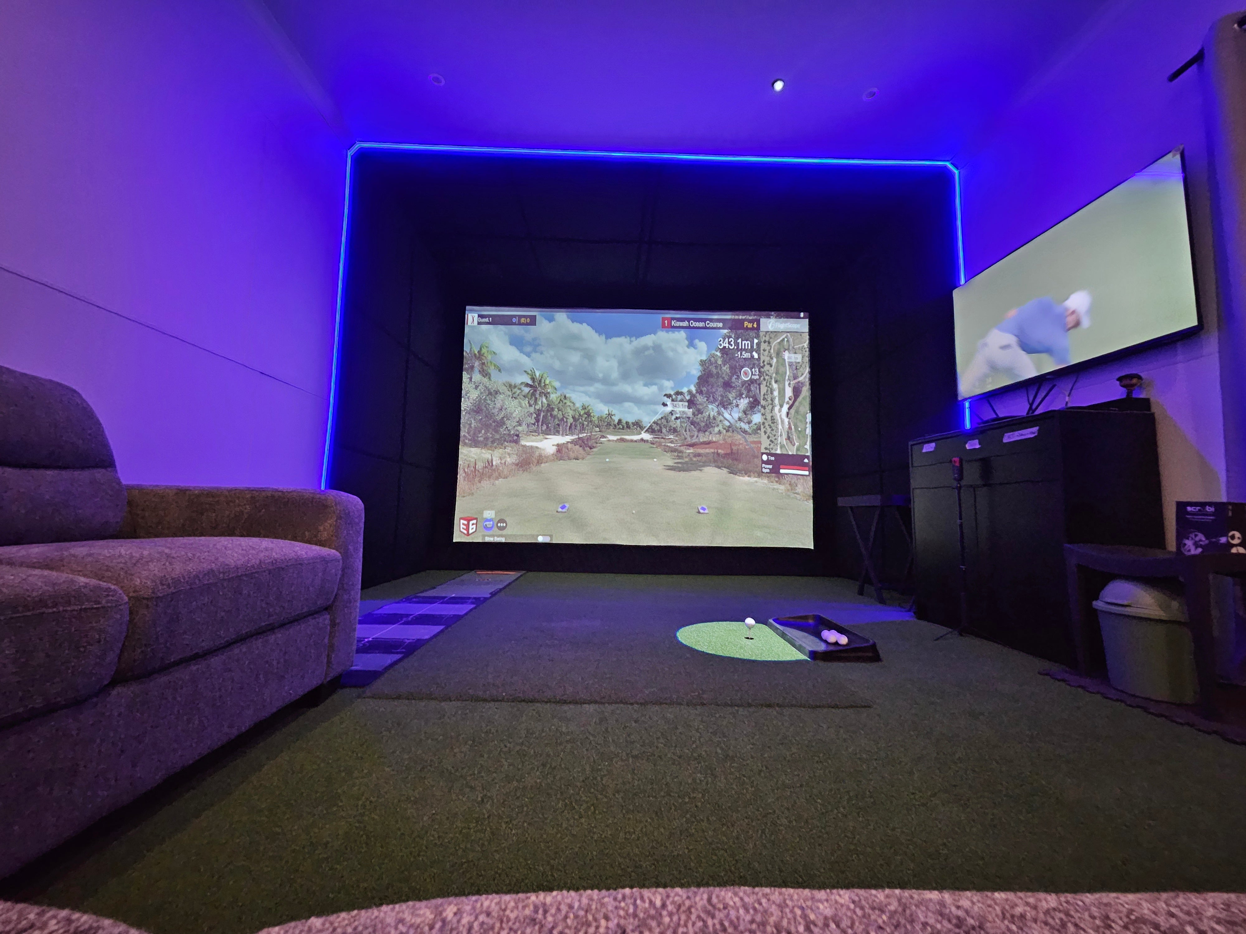 Home golf simulator bay with blue ambient LED lighting, plush seating area, and side monitoring display for data tracking.