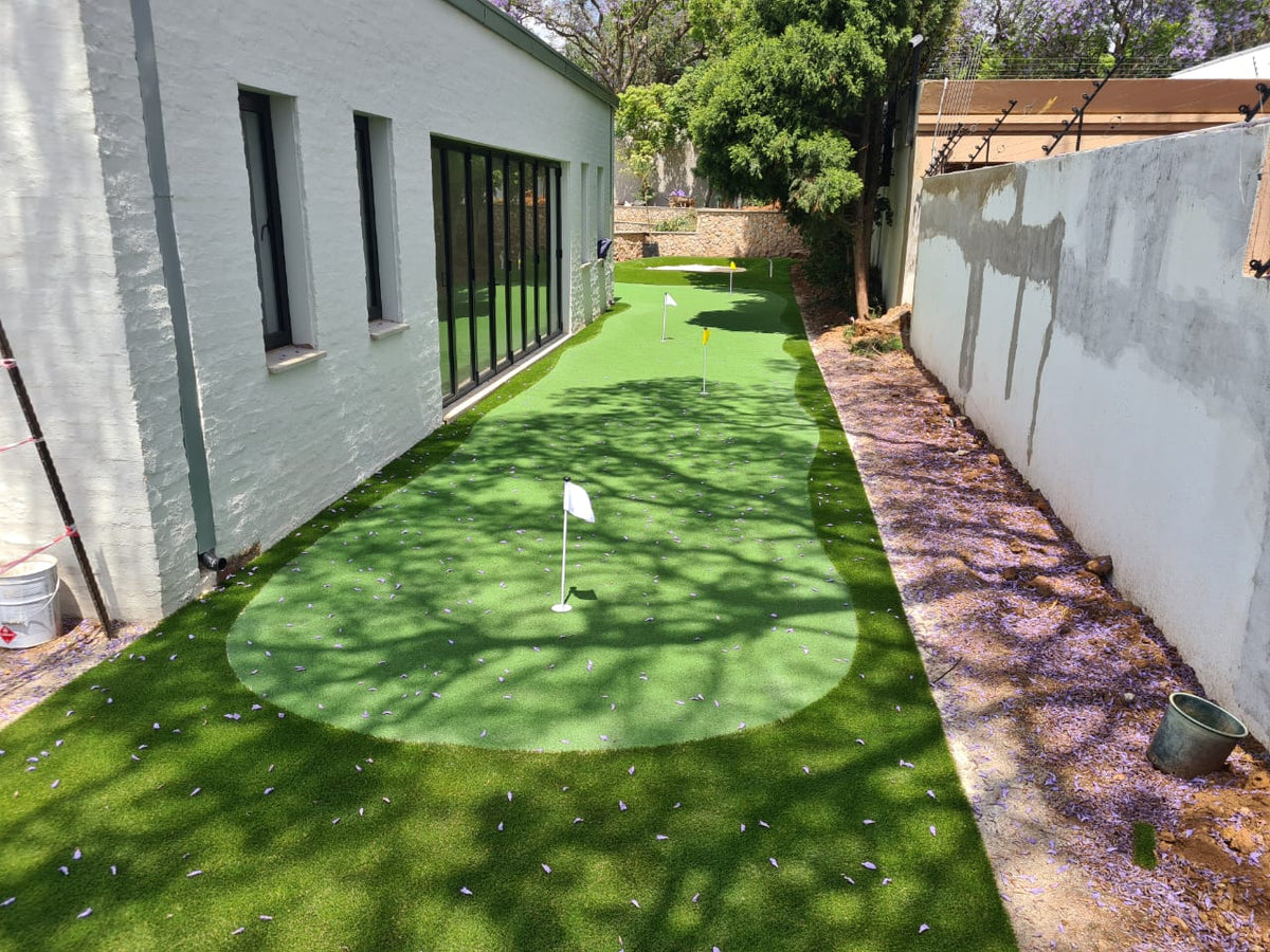Professional outdoor synthetic putting green with an integrated practice sand bunker built into a lush backyard garden.