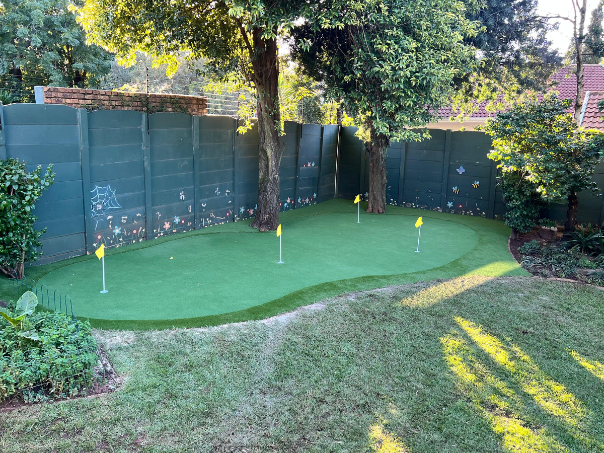 Residential outdoor putting green with yellow target flags installed in a shaded garden corner alongside boundary fencing.